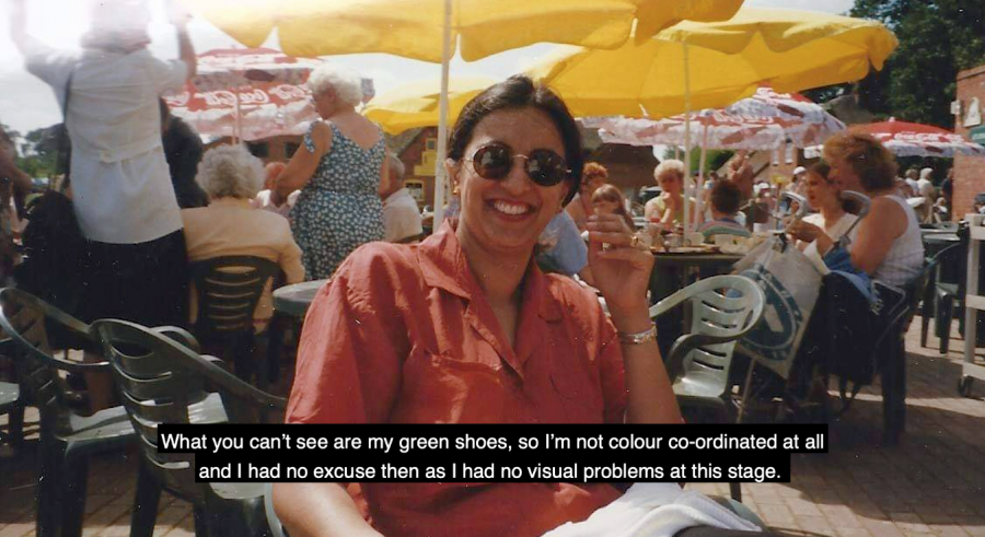 At the centre of this image is an Asian woman in her thirties seated outdoors at a café; she’s smiling and wearing sunglasses and a red shirt. Behind her are tables with yellow umbrellas shading groups of people who are socialising on a sunny day.