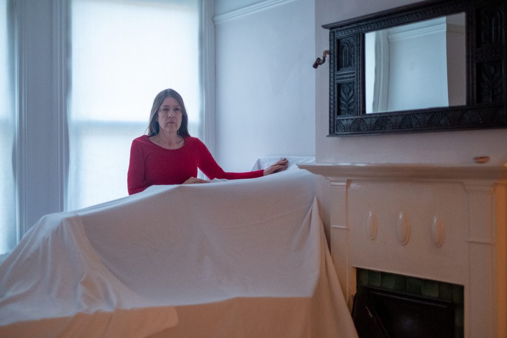 In a bright room a woman in a red top stands behind an item of furniture covered by a white sheet