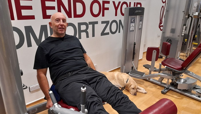 An SVI members works out on one of the weights machines at the gym. His guide dog rests on the floor nearby.