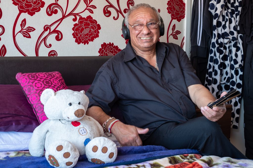 A middle-aged British Asian man, listening to music through headphones, is seated on a bed next to a white teddy bear.