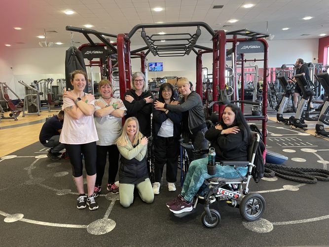 SVI women and friends at the gym. The women are crossing their arms to symbolose embracing equity, this year's theme. One woman is a wheelchair user.
