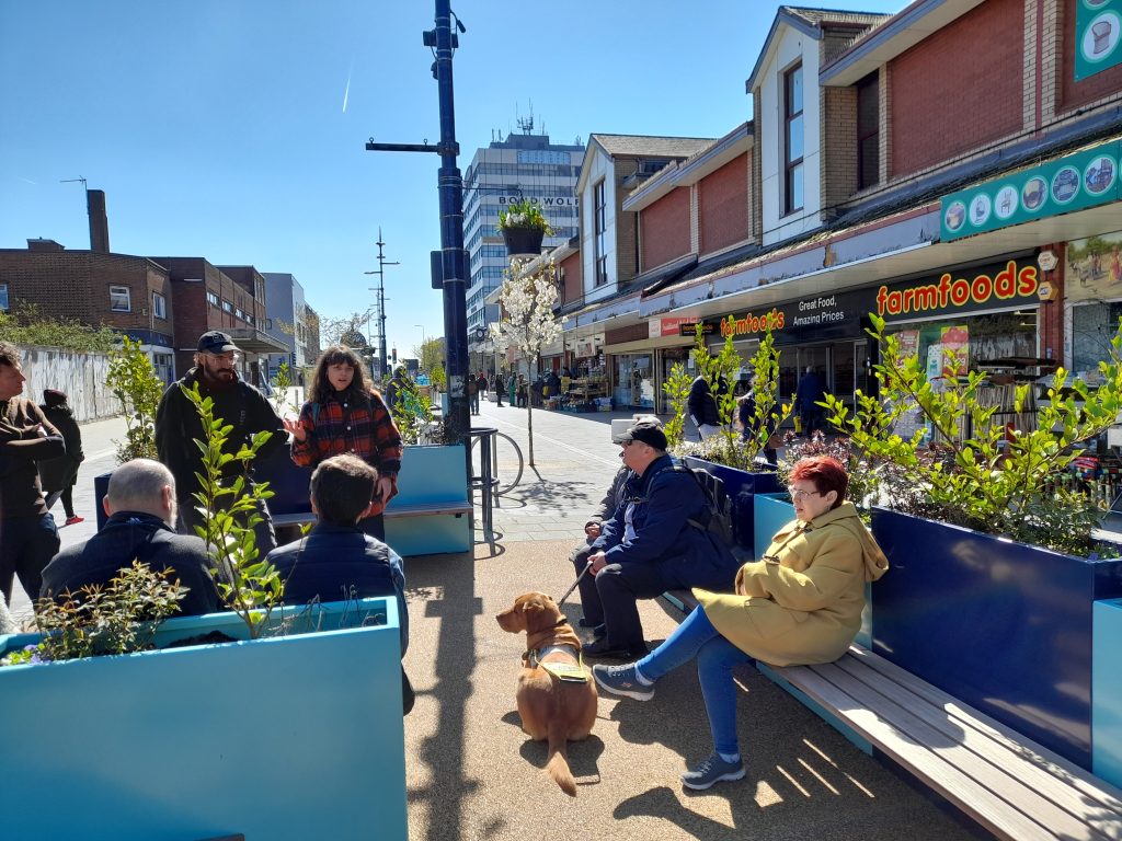 Becky from Multistory introduces the programme. SVI members and a guide dog sit listening in one of the new mini park seating areas on the High Street. The Farmfoods shop is in the background.