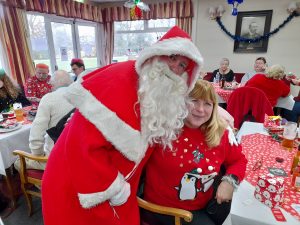 Santa stands with his arm round the back of a seated SVI member, as they pose for a photo. Other members chat and eat in the background.