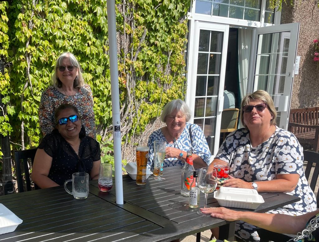 Lots of happy faces at a table with an umbrella on the veranda. There's ivy on the wall in the background. The four ladies have drinks and a takeaway cake box.