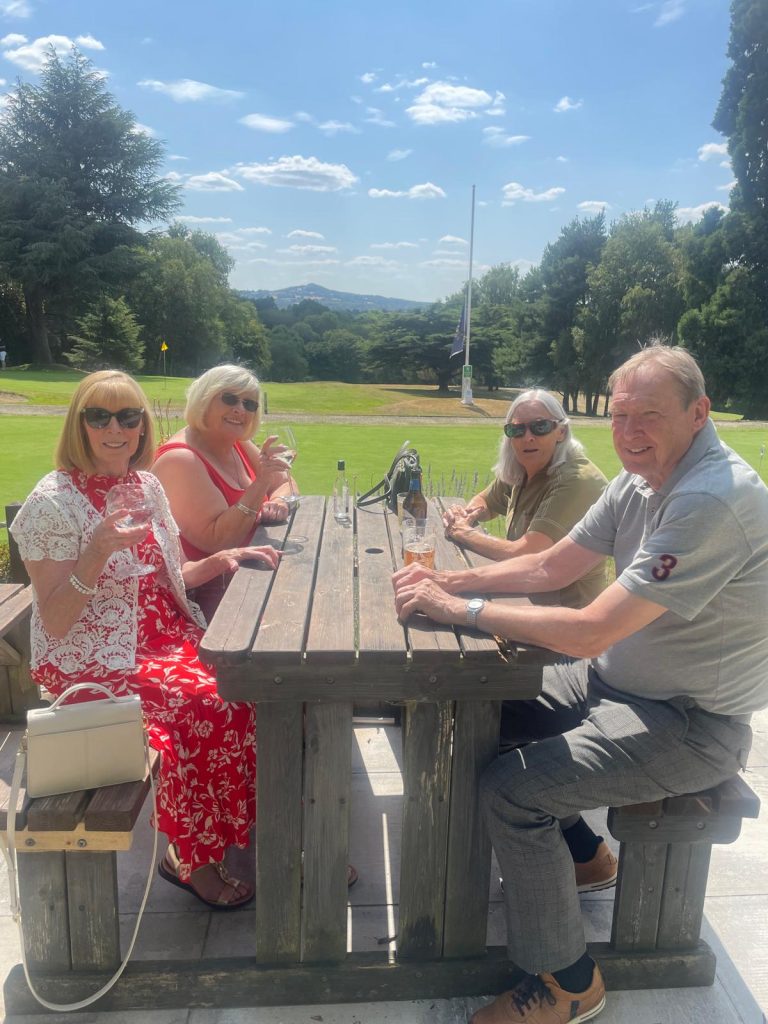 Four SVI members sit at a wooden bench, enjoying a drink with a view of the golf course. The sun is shining and the ladies are wearing summer dresses.