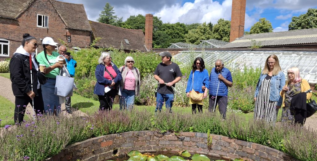 A group of SVI members stand around a lily pond listening to the head gardener describing the plants in and around it.