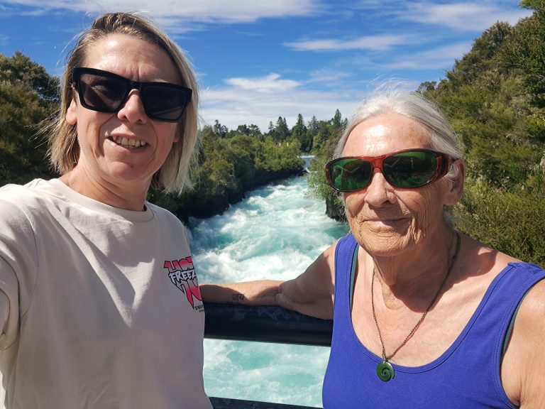 Carrie and her Mum take a selfie standing on a bridge with a rapidly flowing river and a forest in the background