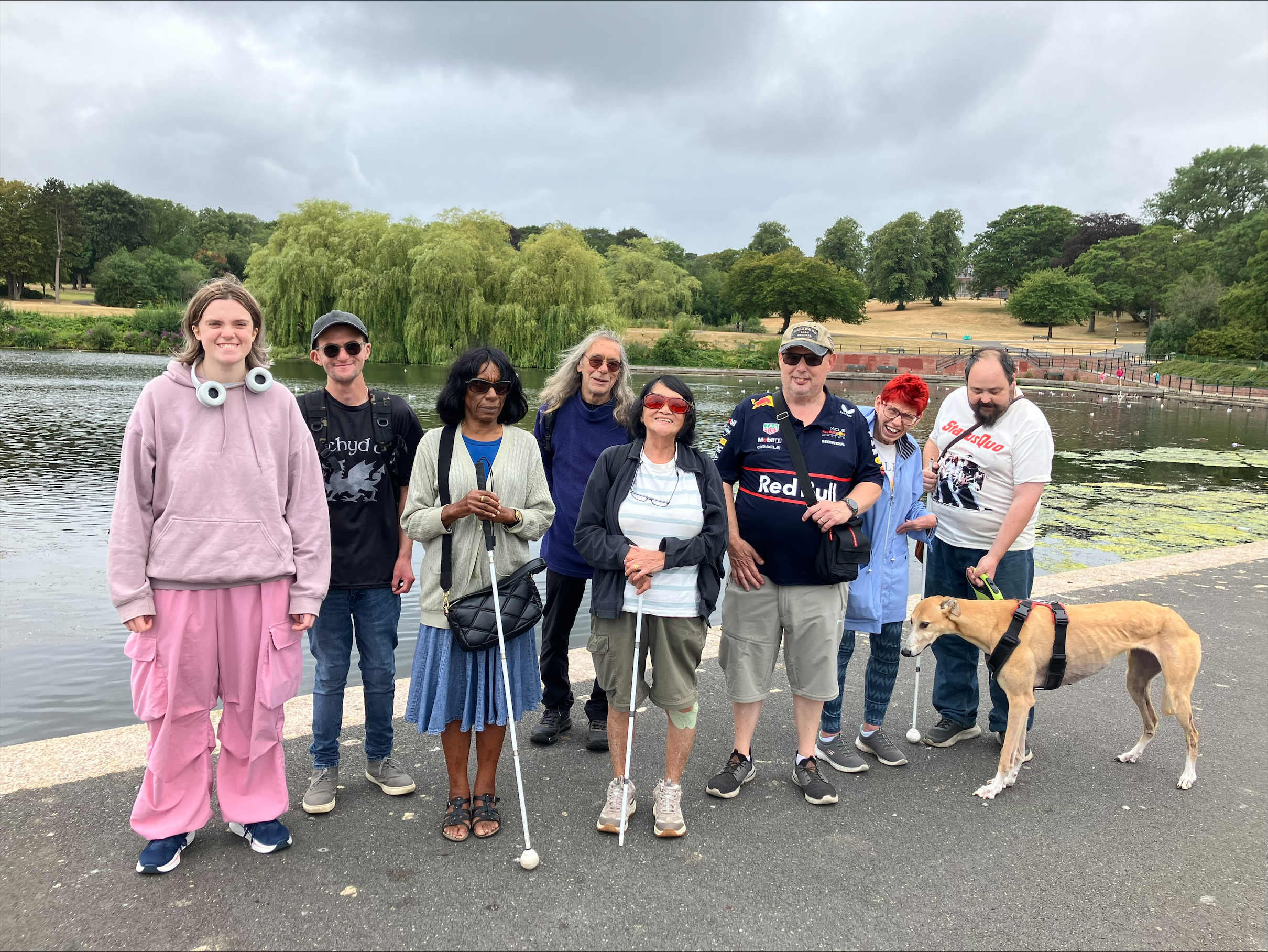 A group of SVI members, some with long canes, stand in a group by a pond in Dartmouth Park. There is a greyhound dog with them, which is a pet not a guide dog.