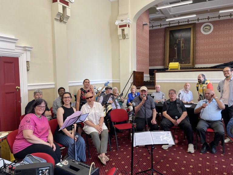 SVI Rocks choir and band performing at Sandwell Visually Impaired's Open Meeting in the Council Chambers at West Bromwich Town Hall. Sixteen performers, led by Rodrigo Orquera, are seated with singers at the front and instrumentalists behind, holding microphones, guitars and percussion instruments.