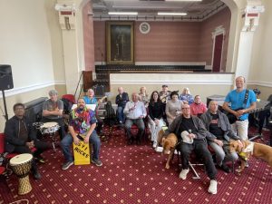 16 SVI Rocks members and two guide dogs sit with guitars, ukuleles, drums and microphones in the chambers of West Bromwich Town Hall, led by band leader Rod